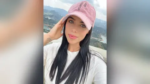 Smiling woman in a white shirt and pink baseball cap enjoying a scenic mountain view outdoors.