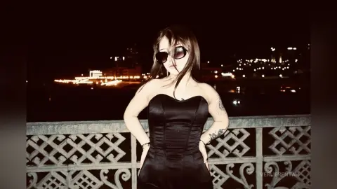 Stylish woman in a black strapless corset posing on a balcony overlooking a glowing city skyline at night.