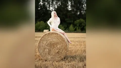 Blonde woman in a white lace dress sitting on a hay bale in a golden field during a dreamy sunset.