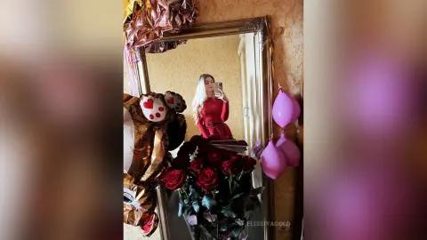Glamorous woman in a red dress taking a selfie in a mirror surrounded by pink balloons and red roses.