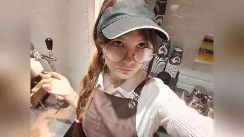 Young woman in a white shirt, apron, and baseball cap working in a modern cafe setting.