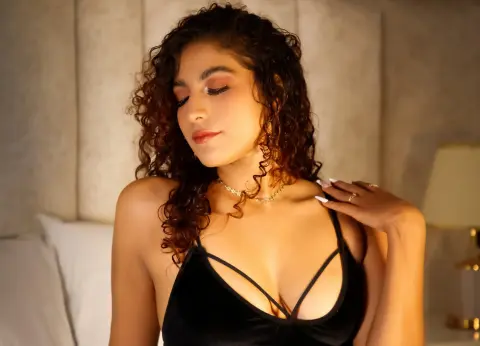Young woman with curly hair in a black velvet top posing in a warm, cozy bedroom setting.