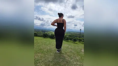 Curvy woman in a black maxi dress standing on a grassy hill under a cloudy sky.