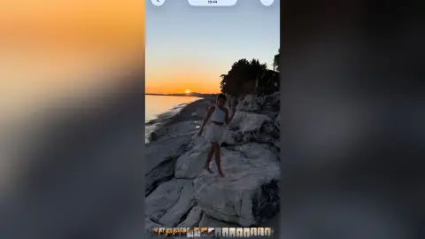 Young woman in a white summer outfit standing on rocky cliffs during a golden sunset at the beach.