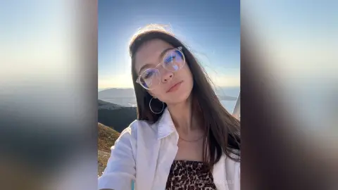 Young woman in glasses and white shirt smiling during a scenic sunset mountain view.