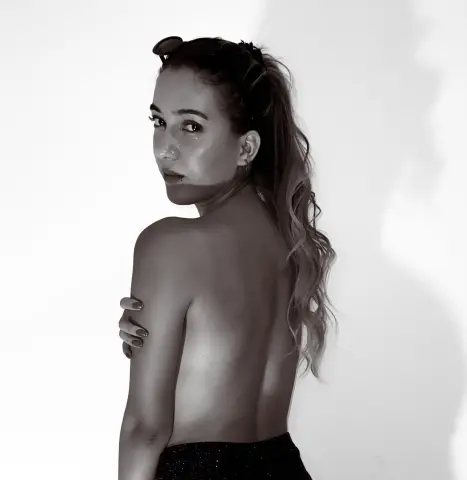 ishabellalopez poses shirtless with her back turned, looking over her shoulder with long wavy hair and sunglasses on her head.