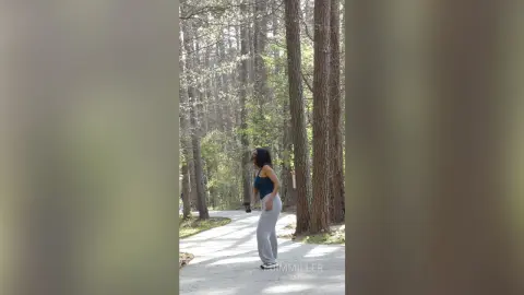 Young woman in a blue tank top and grey sweatpants walking through a peaceful, sunlit forest path.