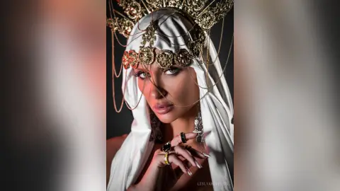 LeslyAnderson wears an ornate gold headpiece and white silk fabric draped over her head and shoulders.