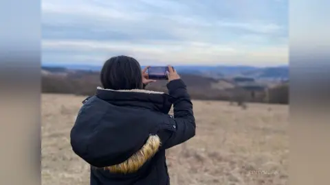 Woman in a black winter jacket taking a photo of a vast landscape view under a cloudy sky.