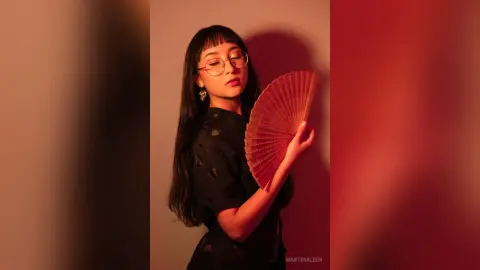 Stylish woman in a black patterned blouse holding a red folding fan against a warm studio background.