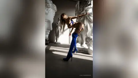 LeslyAnderson arches her back while wearing a blue bikini and matching thigh-high boots against a white statue background.