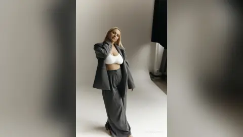 Blonde woman in a grey oversized suit and white bralette posing confidently in a minimalist studio setting.