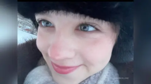 AmberLune smiles while wearing a black furry hat and a light grey scarf against a snowy outdoor background.
