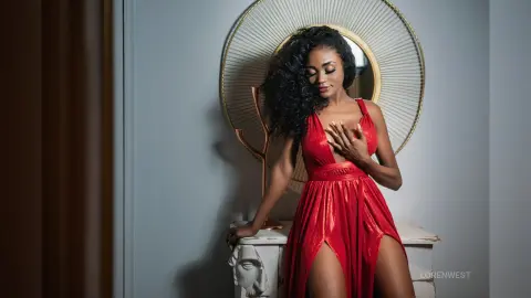 Elegant woman in a glamorous red evening gown posing by a gold mirror in a sophisticated indoor setting.
