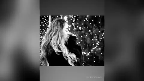 TiffanyAddams poses in profile with her long, wavy hair falling over her shoulder against a background of soft, glowing bokeh lights.