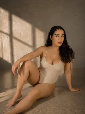 paulinaa_rosee poses sitting on the floor in a light tan bodysuit, showing her long dark curly hair and cleavage against a neutral wall.