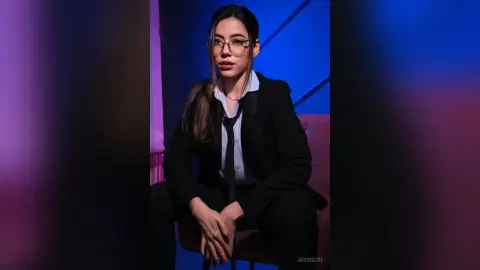 JessieLiu wears a black suit and tie with a white shirt and glasses while sitting in a dimly lit room.