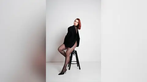 AryaSin poses in a black blazer and sheer black tights while sitting on a stool against a white background.