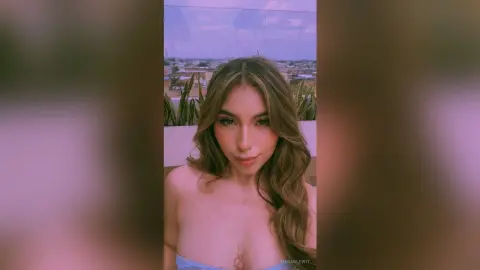 MeganLewit poses with long wavy hair, wearing a light blue bra that shows her cleavage and bare shoulders against an outdoor background.