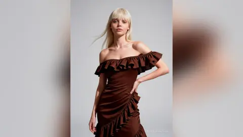 FaviolaKezele poses in a brown off-the-shoulder ruffled dress with her hand on her hip against a plain grey background.