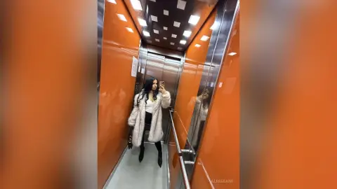 AlmaKane — Stylish woman in a cozy white faux fur coat taking a mirror selfie in a modern orange elevator.