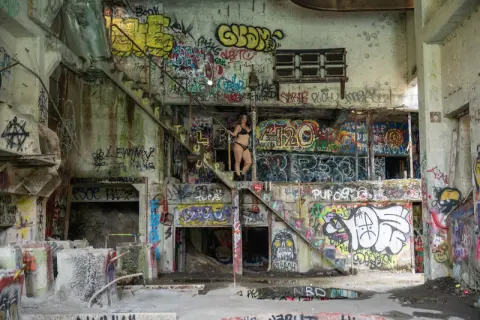 Woman in black bikini standing in a gritty, graffiti-covered abandoned building with urban decay vibes.