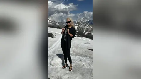 Stylish woman in black outfit and sunglasses standing on a snowy mountain landscape under a cloudy sky.
