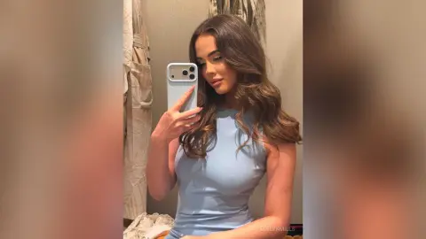 Young woman in a light blue sleeveless top taking a mirror selfie in a cozy indoor setting.