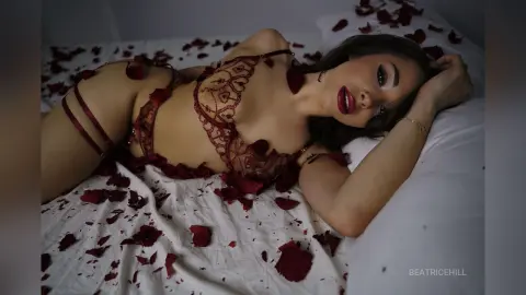 Young woman in red lace lingerie lying on white bedding surrounded by scattered rose petals in a romantic mood.