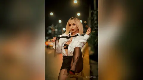 Blonde woman in a white shirt and black skirt posing confidently on a city street at night.