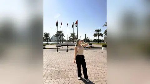 Blonde woman in a beige tank top and black trousers standing in a sunny plaza with flags under a clear sky.