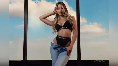MiaGuerra poses in a black Undergold bra and matching undies with her light brown hair down, wearing light blue jeans against a blue sky background.
