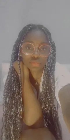 Young woman with long dark braids and pink glasses wearing a white top in a soft, moody indoor setting.