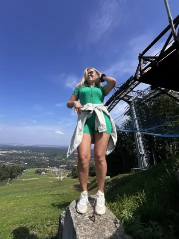 NikaZen stands on a rock outdoors in a green top and shorts, looking up at the sky with a light denim jacket tied around her waist.
