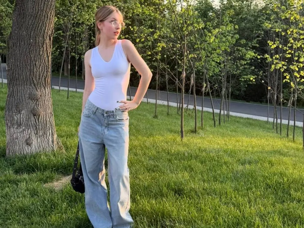 Young woman in white tank top and light blue jeans standing in a sunny green park meadow.