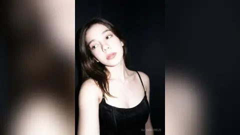 Young woman in a black satin camisole top posing with a moody, cinematic look against a dark background.
