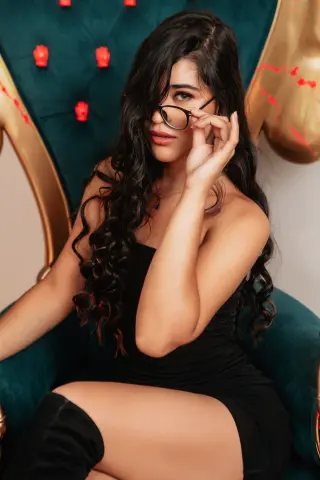 Elegant woman in a black dress and glasses sitting on a teal velvet throne in a moody, luxurious setting.