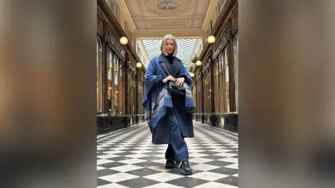 DaisyLin stands in a grand hallway wearing a blue wrap, black top, and dark jeans.