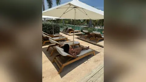 Relaxed woman in swimwear lounging on a sunbed under a white umbrella by a tropical resort swimming pool.