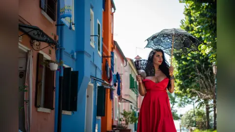 MissyJolie stands in a red dress holding a black lace parasol against a colorful street background.