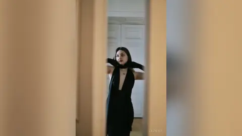 Elegant woman in a black sleeveless dress and scarf posing in a minimalist indoor setting with a moody vibe.