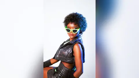 Stylish Black woman in black leather outfit and green sunglasses posing against a bright studio background.