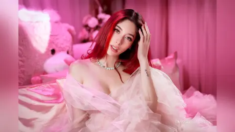 Red-haired woman in a dreamy pink ruffled gown sitting on a bed in a soft romantic setting.