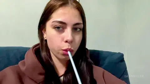 Young woman in a brown hoodie sipping a drink through a straw while sitting on a blue sofa.