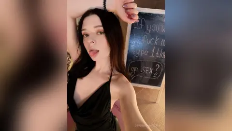 Young woman in a black satin dress posing playfully in front of a chalkboard sign in a cozy room.