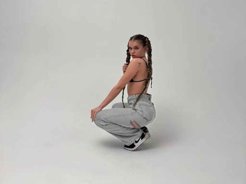 SamaraAdam squats in light wash denim jeans and a black bikini top, showing off her long dark braids against a plain white background.
