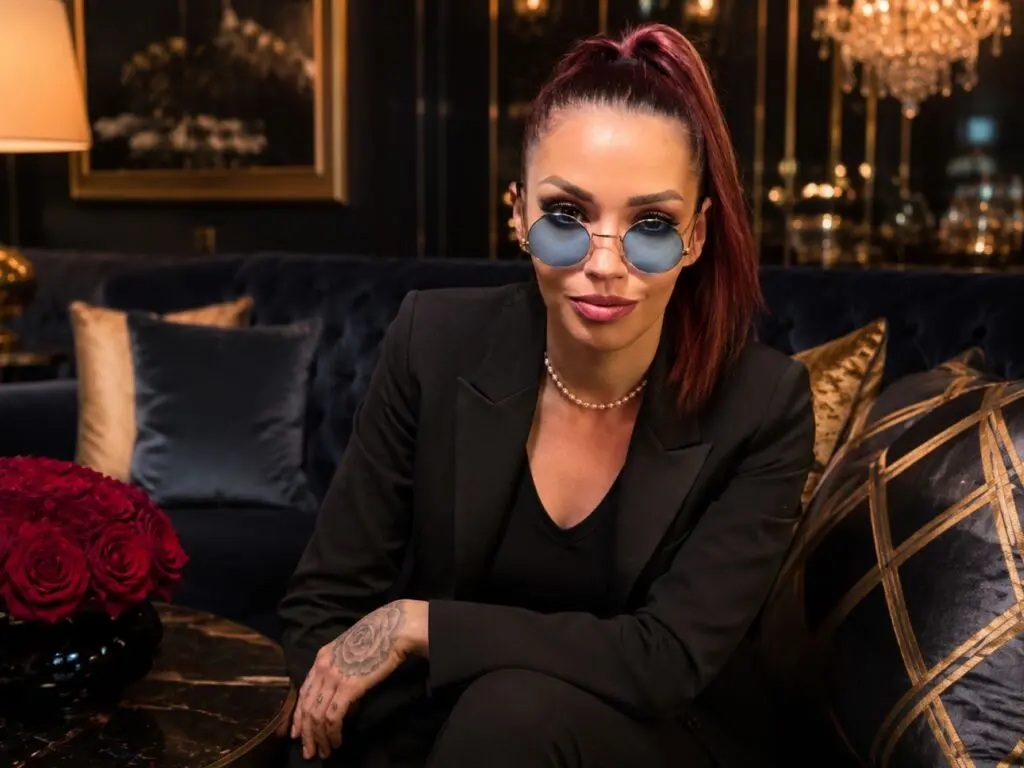 Stylish woman in a black blazer and blue sunglasses sitting on a velvet sofa in a luxury lounge setting.