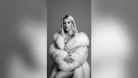 JessycaPage poses in a fluffy white fur coat with her blonde hair down against a plain background.