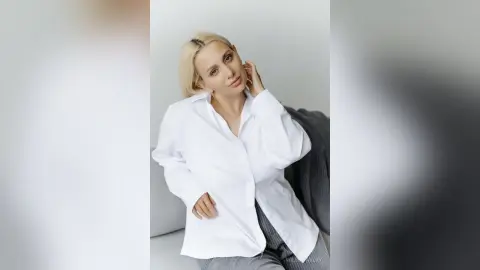 Blonde woman in a white oversized button-down shirt posing elegantly in a bright minimalist studio.