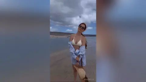 Stylish woman in a white bikini and blue shirt relaxing on a cloudy beach with a straw hat.